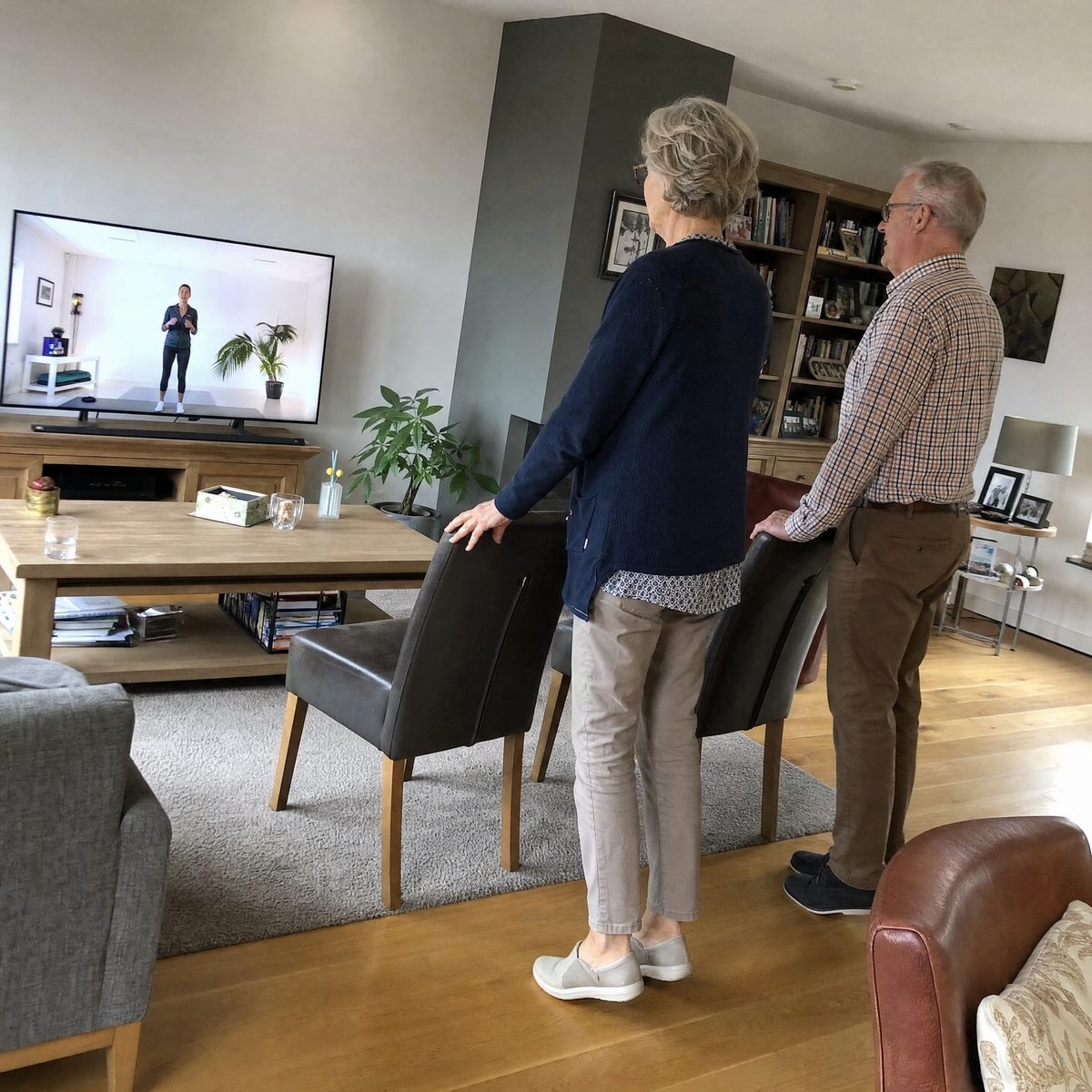 Een stel volgt samen in de woonkamer een les op de televisie, met de handen op een stoelleuning als steun